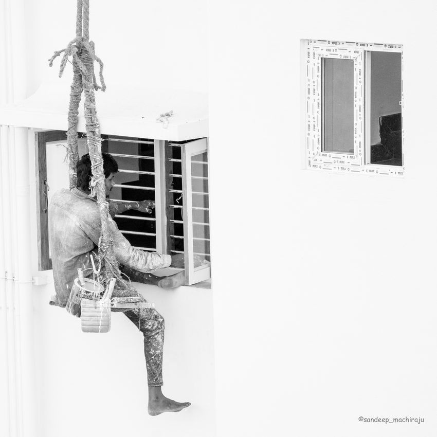 A painter suspended beside a building, at work on a window, in black and white.