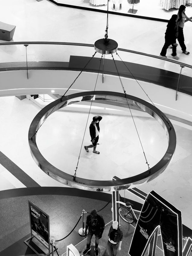 People moving through a shopping centre beneath a large circular light, in black and white.