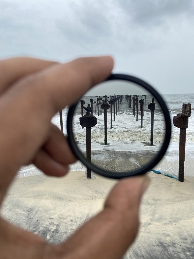 A hand holding a circular camera filter in front of a line of old pier posts extending into the sea.